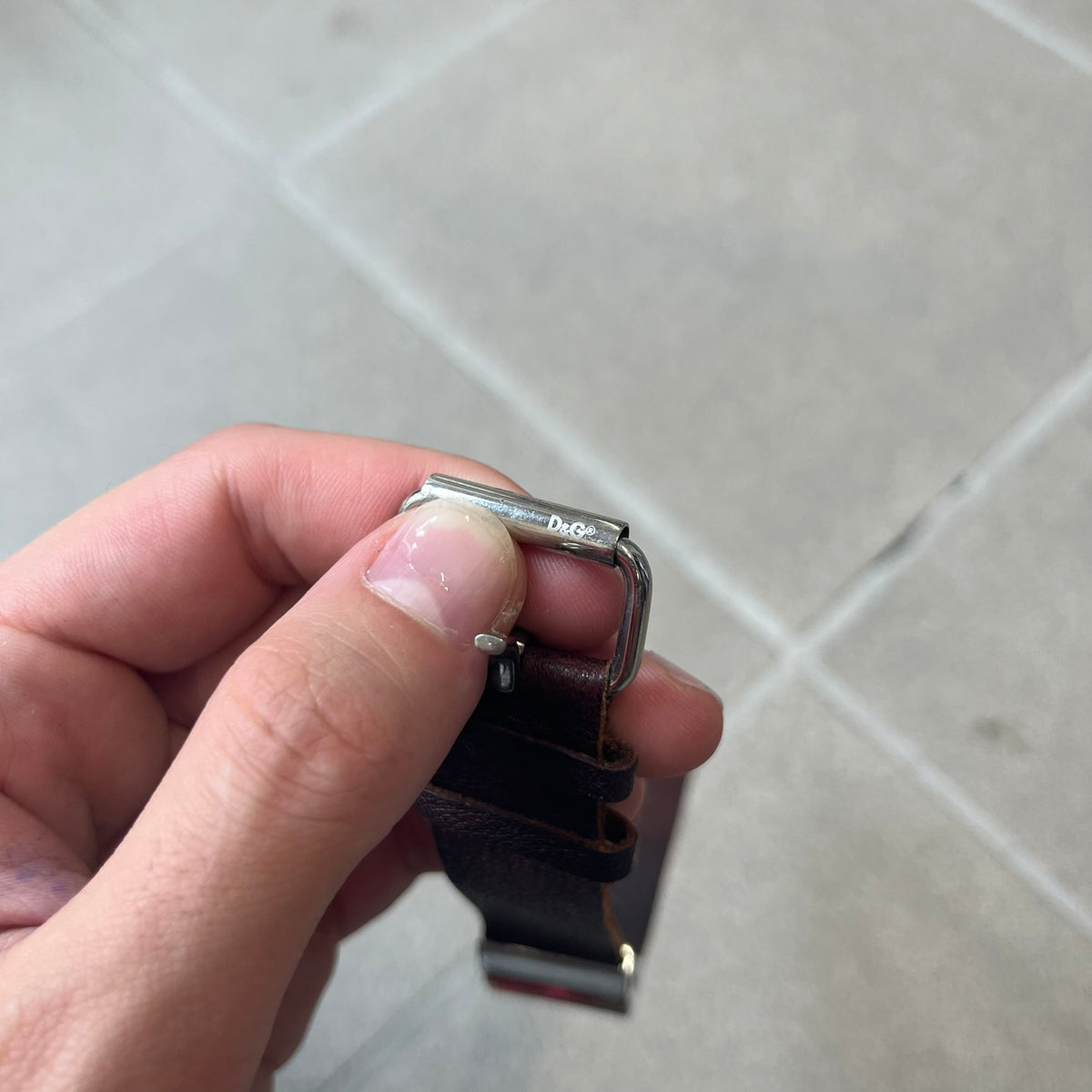 Dolce & Gabbana 2000's Brown Leather Bracelet features brown leather straps with an adjustable fit and a stainless steel central plaque. The plaque has a raised wavy texture on the front and a D&G logo underneath, while the leather straps have small D&G and “Genuine Leather” tags.
