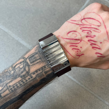 Dolce & Gabbana 2000's Brown Leather Bracelet features brown leather straps with an adjustable fit and a stainless steel central plaque. The plaque has a raised wavy texture on the front and a D&G logo underneath, while the leather straps have small D&G and “Genuine Leather” tags.
