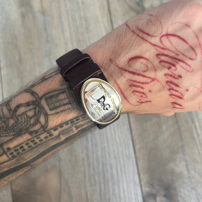 Dolce &amp; Gabbana Champagne Leather Bracelet DJ1097 is a brown leather bracelet designed to resemble a miniature belt, featuring multiple adjustment holes. The silver-tone metal plate is shaped like a D&amp;G champagne label and displays a black D&amp;G logo, along with engraved “Glamorous Champagne 1994 Alc. 12% by Vol.” text. The back of the plate features a large, shallow engraved D&amp;G logo.