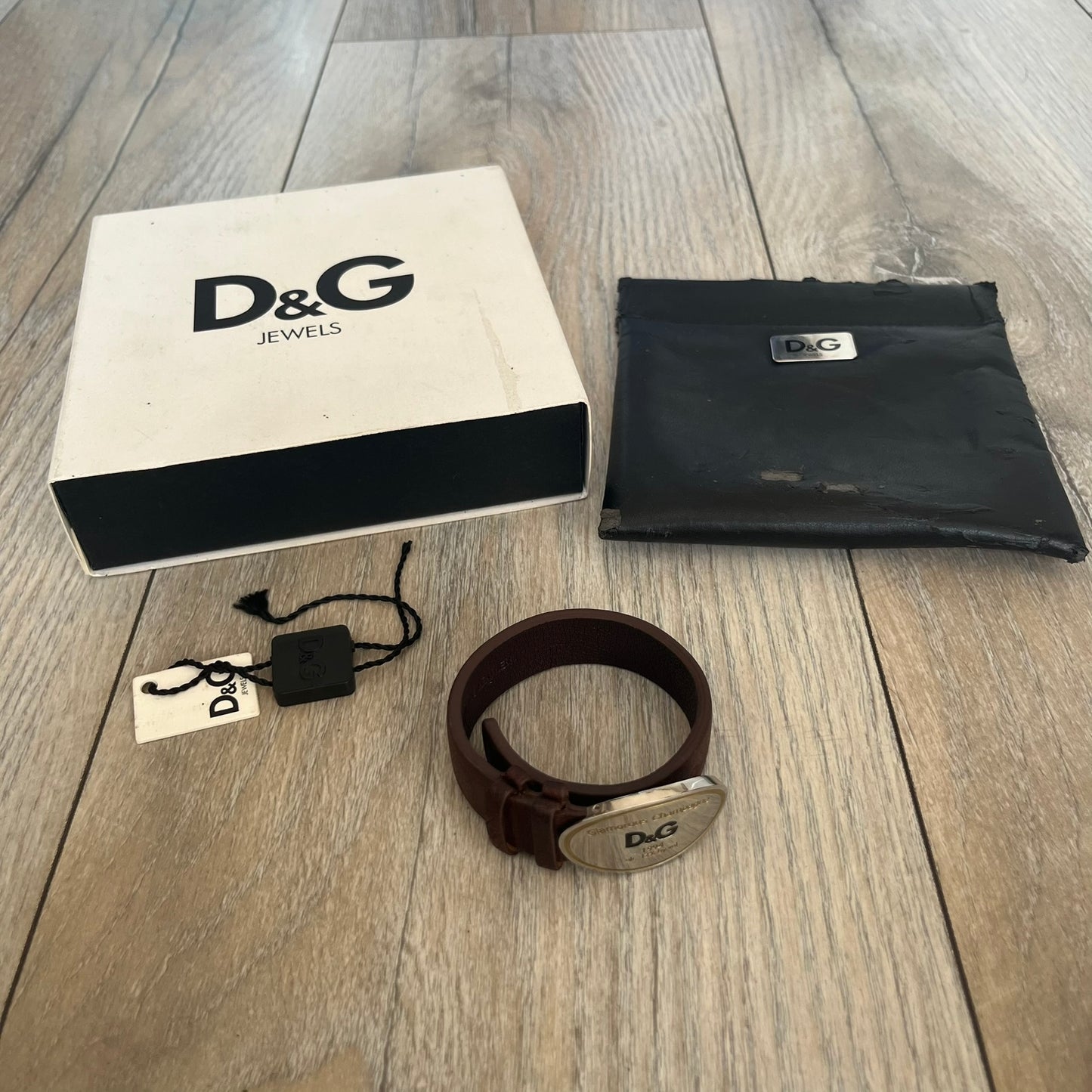 Dolce &amp; Gabbana Champagne Leather Bracelet DJ1097 is a brown leather bracelet designed to resemble a miniature belt, featuring multiple adjustment holes. The silver-tone metal plate is shaped like a D&amp;G champagne label and displays a black D&amp;G logo, along with engraved “Glamorous Champagne 1994 Alc. 12% by Vol.” text. The back of the plate features a large, shallow engraved D&amp;G logo.