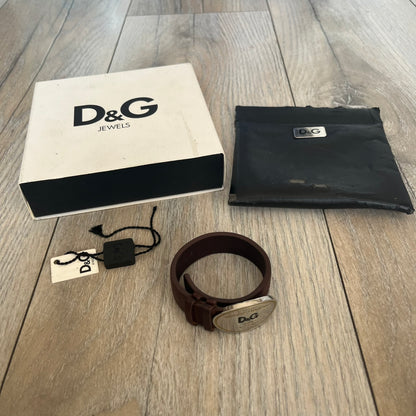 Dolce &amp; Gabbana Champagne Leather Bracelet DJ1097 is a brown leather bracelet designed to resemble a miniature belt, featuring multiple adjustment holes. The silver-tone metal plate is shaped like a D&amp;G champagne label and displays a black D&amp;G logo, along with engraved “Glamorous Champagne 1994 Alc. 12% by Vol.” text. The back of the plate features a large, shallow engraved D&amp;G logo.