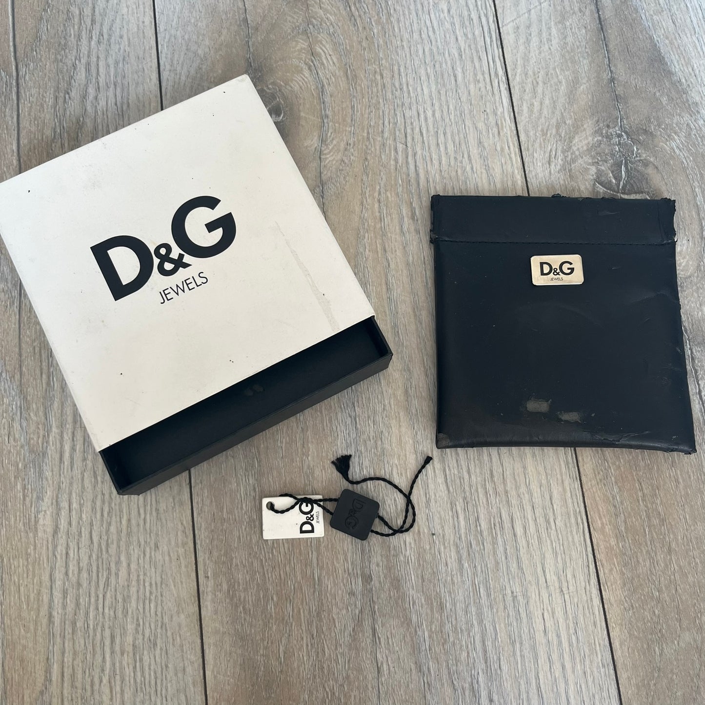 Dolce &amp; Gabbana Champagne Leather Bracelet DJ1097 is a brown leather bracelet designed to resemble a miniature belt, featuring multiple adjustment holes. The silver-tone metal plate is shaped like a D&amp;G champagne label and displays a black D&amp;G logo, along with engraved “Glamorous Champagne 1994 Alc. 12% by Vol.” text. The back of the plate features a large, shallow engraved D&amp;G logo.