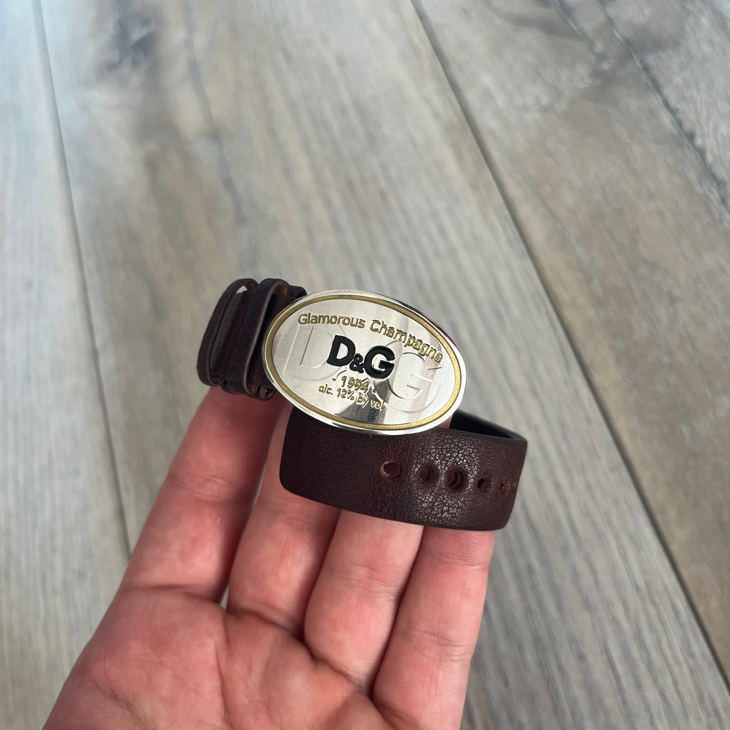 Dolce &amp; Gabbana Champagne Leather Bracelet DJ1097 is a brown leather bracelet designed to resemble a miniature belt, featuring multiple adjustment holes. The silver-tone metal plate is shaped like a D&amp;G champagne label and displays a black D&amp;G logo, along with engraved “Glamorous Champagne 1994 Alc. 12% by Vol.” text. The back of the plate features a large, shallow engraved D&amp;G logo.