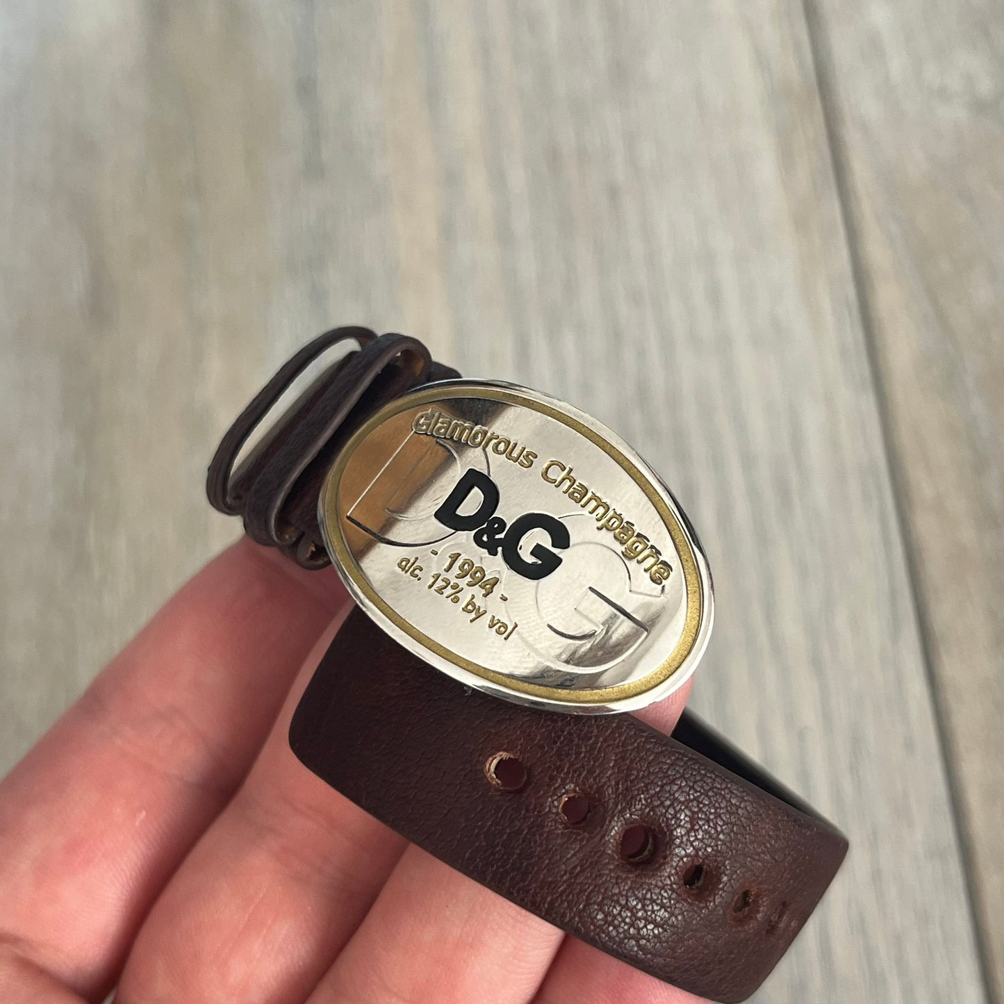 Dolce &amp; Gabbana Champagne Leather Bracelet DJ1097 is a brown leather bracelet designed to resemble a miniature belt, featuring multiple adjustment holes. The silver-tone metal plate is shaped like a D&amp;G champagne label and displays a black D&amp;G logo, along with engraved “Glamorous Champagne 1994 Alc. 12% by Vol.” text. The back of the plate features a large, shallow engraved D&amp;G logo.