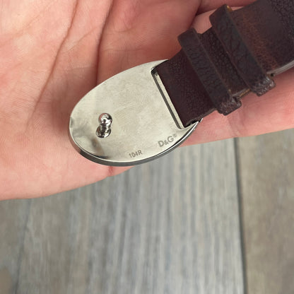 Dolce &amp; Gabbana Champagne Leather Bracelet DJ1097 is a brown leather bracelet designed to resemble a miniature belt, featuring multiple adjustment holes. The silver-tone metal plate is shaped like a D&amp;G champagne label and displays a black D&amp;G logo, along with engraved “Glamorous Champagne 1994 Alc. 12% by Vol.” text. The back of the plate features a large, shallow engraved D&amp;G logo.