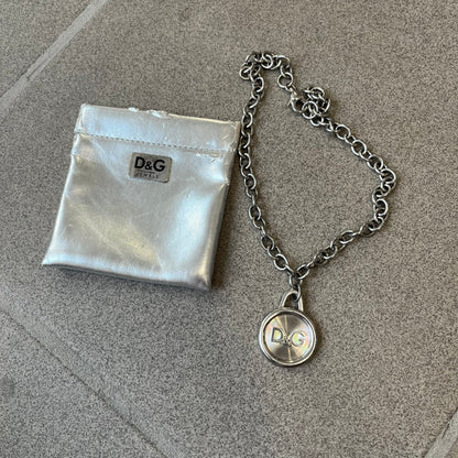 Dolce & Gabbana 2000's Circular Pendant Necklace features a circular pendant with a flat stainless steel base, raised outer detailing, and a raised D&G logo in the center. The pendant has a small triangular loop at the top connecting it to the chain. Comes with a dust bag.