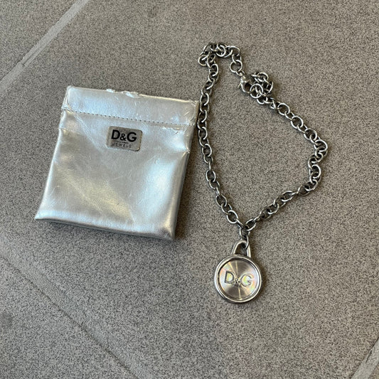 Dolce & Gabbana 2000's Circular Pendant Necklace features a circular pendant with a flat stainless steel base, raised outer detailing, and a raised D&G logo in the center. The pendant has a small triangular loop at the top connecting it to the chain. Comes with a dust bag.