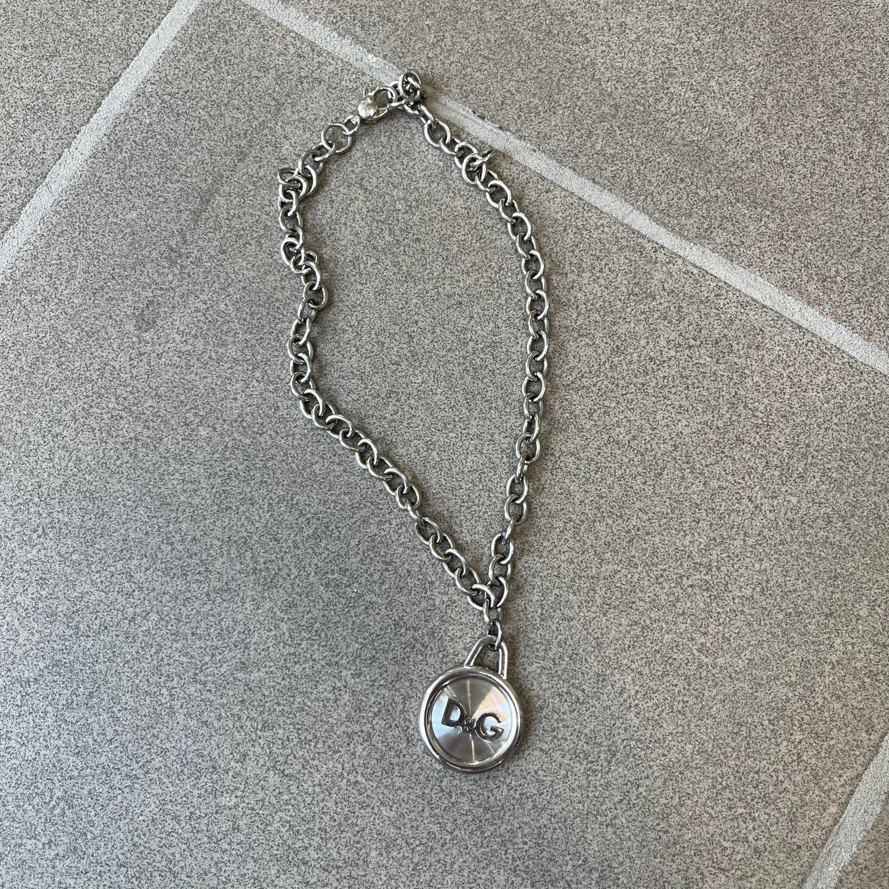 Dolce & Gabbana 2000's Circular Pendant Necklace features a circular pendant with a flat stainless steel base, raised outer detailing, and a raised D&G logo in the center. The pendant has a small triangular loop at the top connecting it to the chain. Comes with a dust bag.