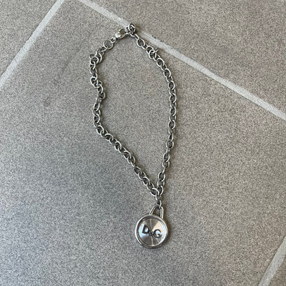 Dolce & Gabbana 2000's Circular Pendant Necklace features a circular pendant with a flat stainless steel base, raised outer detailing, and a raised D&G logo in the center. The pendant has a small triangular loop at the top connecting it to the chain. Comes with a dust bag.