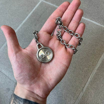 Dolce & Gabbana 2000's Circular Pendant Necklace features a circular pendant with a flat stainless steel base, raised outer detailing, and a raised D&G logo in the center. The pendant has a small triangular loop at the top connecting it to the chain. Comes with a dust bag.