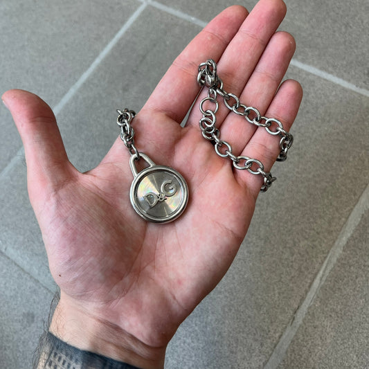 Dolce & Gabbana 2000's Circular Pendant Necklace features a circular pendant with a flat stainless steel base, raised outer detailing, and a raised D&G logo in the center. The pendant has a small triangular loop at the top connecting it to the chain. Comes with a dust bag.