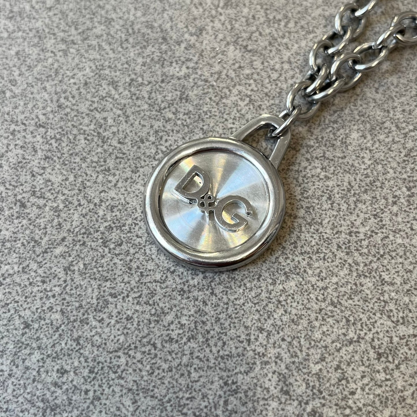 Dolce & Gabbana 2000's Circular Pendant Necklace features a circular pendant with a flat stainless steel base, raised outer detailing, and a raised D&G logo in the center. The pendant has a small triangular loop at the top connecting it to the chain. Comes with a dust bag.