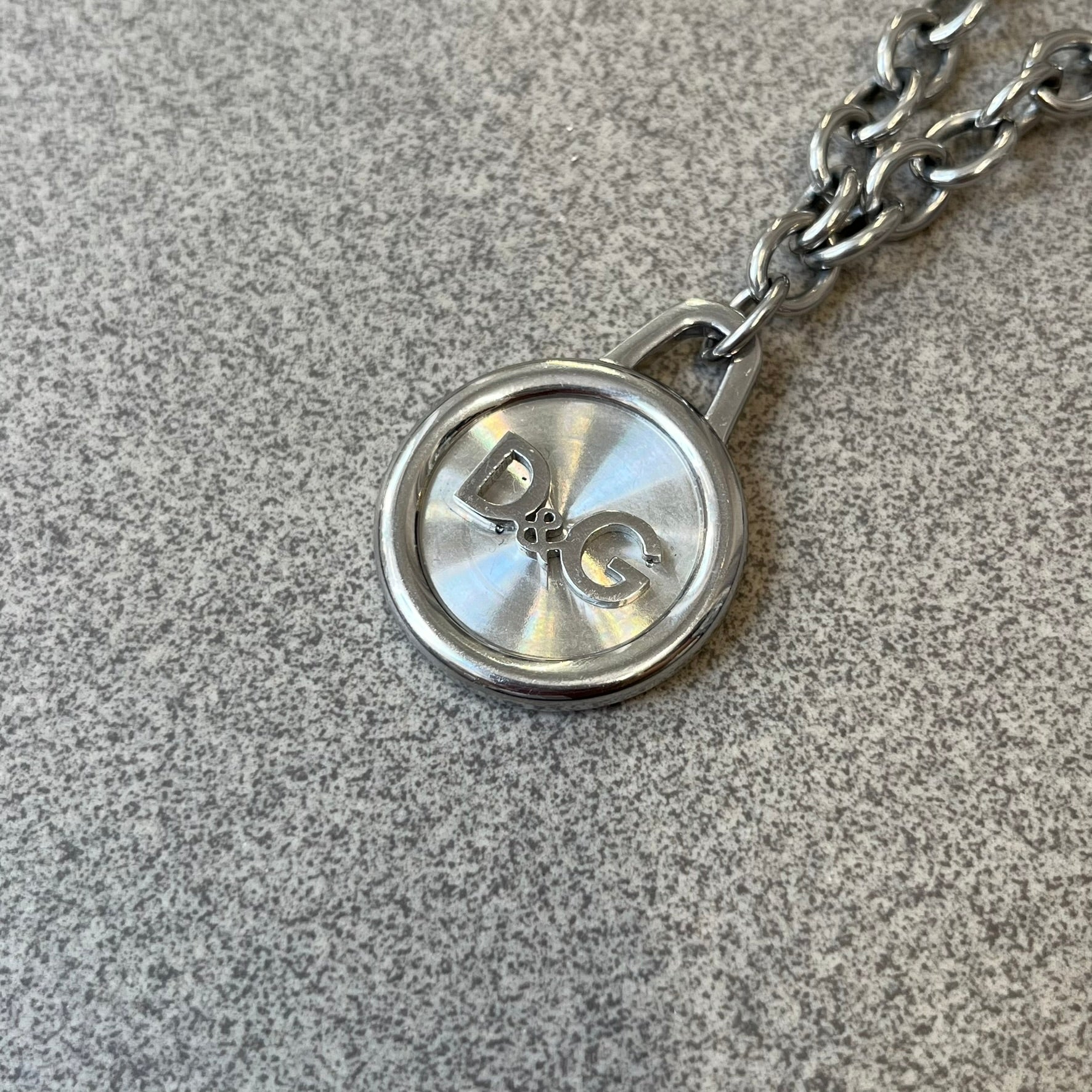 Dolce & Gabbana 2000's Circular Pendant Necklace features a circular pendant with a flat stainless steel base, raised outer detailing, and a raised D&G logo in the center. The pendant has a small triangular loop at the top connecting it to the chain. Comes with a dust bag.