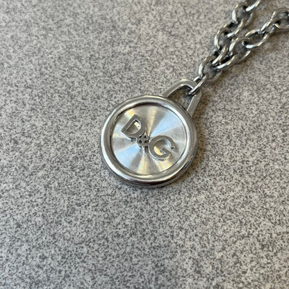 Dolce & Gabbana 2000's Circular Pendant Necklace features a circular pendant with a flat stainless steel base, raised outer detailing, and a raised D&G logo in the center. The pendant has a small triangular loop at the top connecting it to the chain. Comes with a dust bag.