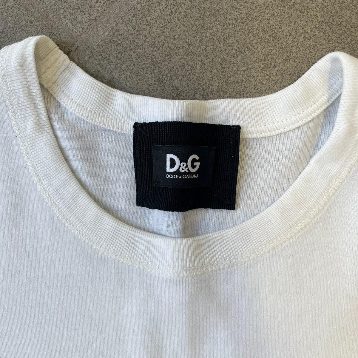 Dolce &amp; Gabbana 2000’s Claudia Schiffer Photo White Tee is a white T-shirt with a photo of naked Claudia Schiffer on the front, covering her chest with her hand, and small “Photo by Mario Testino” text below it. The upper back has “D&amp;G 18 La Lune” text. The shirt is constructed inside-out style, with the stitching visible along the sides.