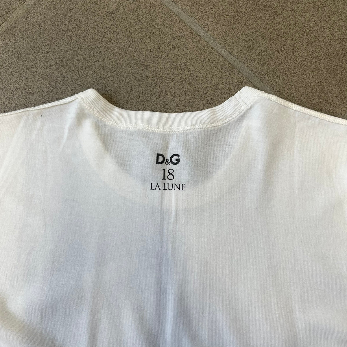 Dolce &amp; Gabbana 2000’s Claudia Schiffer Photo White Tee is a white T-shirt with a photo of naked Claudia Schiffer on the front, covering her chest with her hand, and small “Photo by Mario Testino” text below it. The upper back has “D&amp;G 18 La Lune” text. The shirt is constructed inside-out style, with the stitching visible along the sides.