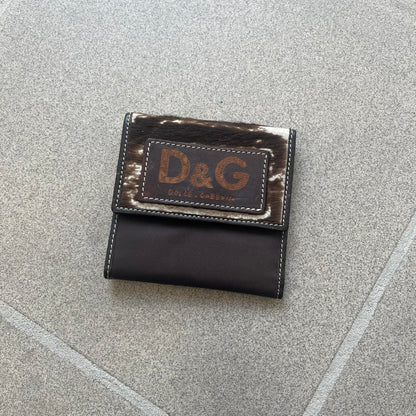Dolce & Gabbana 2000s Cow-Print Wallet features a brown leather construction with a cow-print pony hair-style panel on the front exterior. A leather patch with a D&G logo is placed over the patterned section. The interior includes multiple compartments, card slots, a top compartment, and a zipped pocket for storage. Made in Italy.

