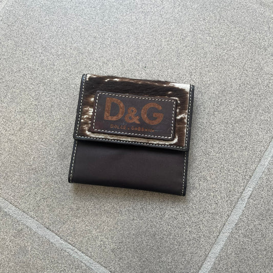 Dolce & Gabbana 2000s Cow-Print Wallet features a brown leather construction with a cow-print pony hair-style panel on the front exterior. A leather patch with a D&G logo is placed over the patterned section. The interior includes multiple compartments, card slots, a top compartment, and a zipped pocket for storage. Made in Italy.
