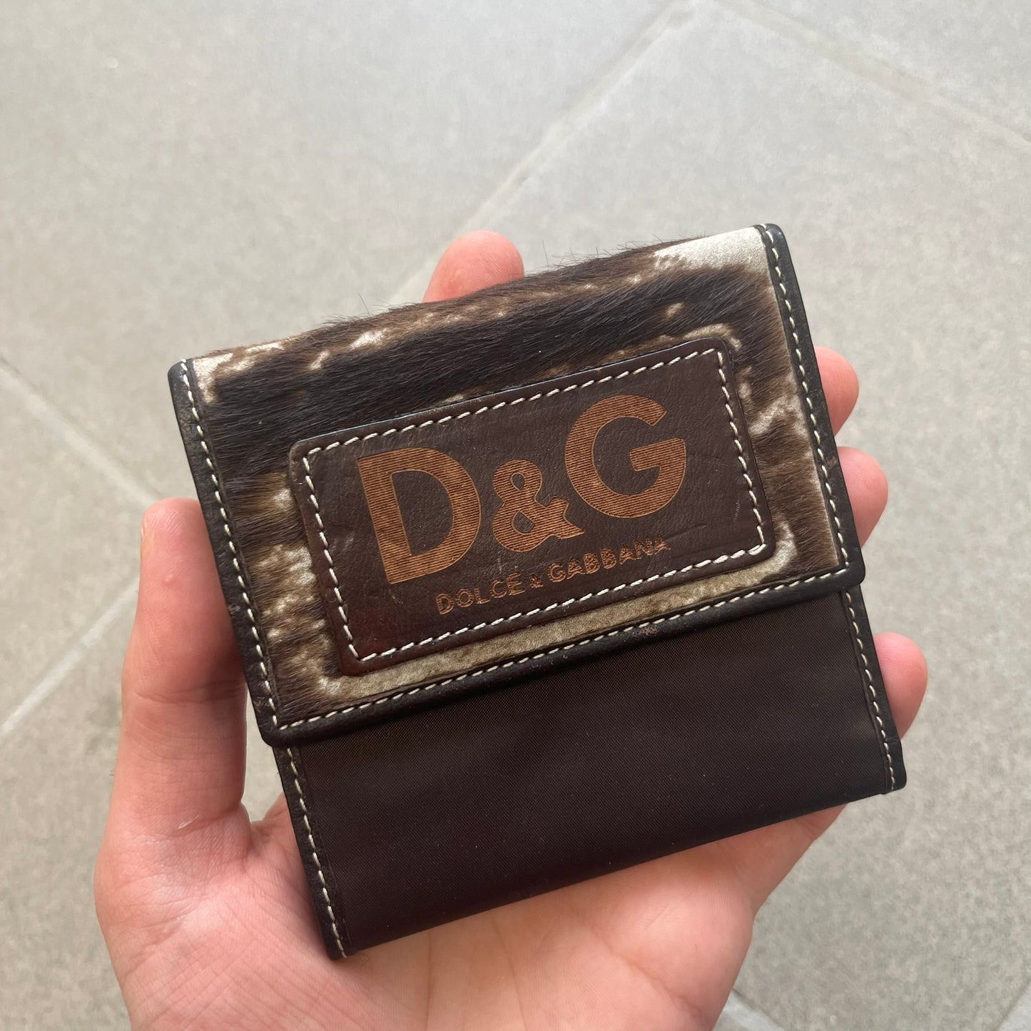 Dolce & Gabbana 2000s Cow-Print Wallet features a brown leather construction with a cow-print pony hair-style panel on the front exterior. A leather patch with a D&G logo is placed over the patterned section. The interior includes multiple compartments, card slots, a top compartment, and a zipped pocket for storage. Made in Italy.
