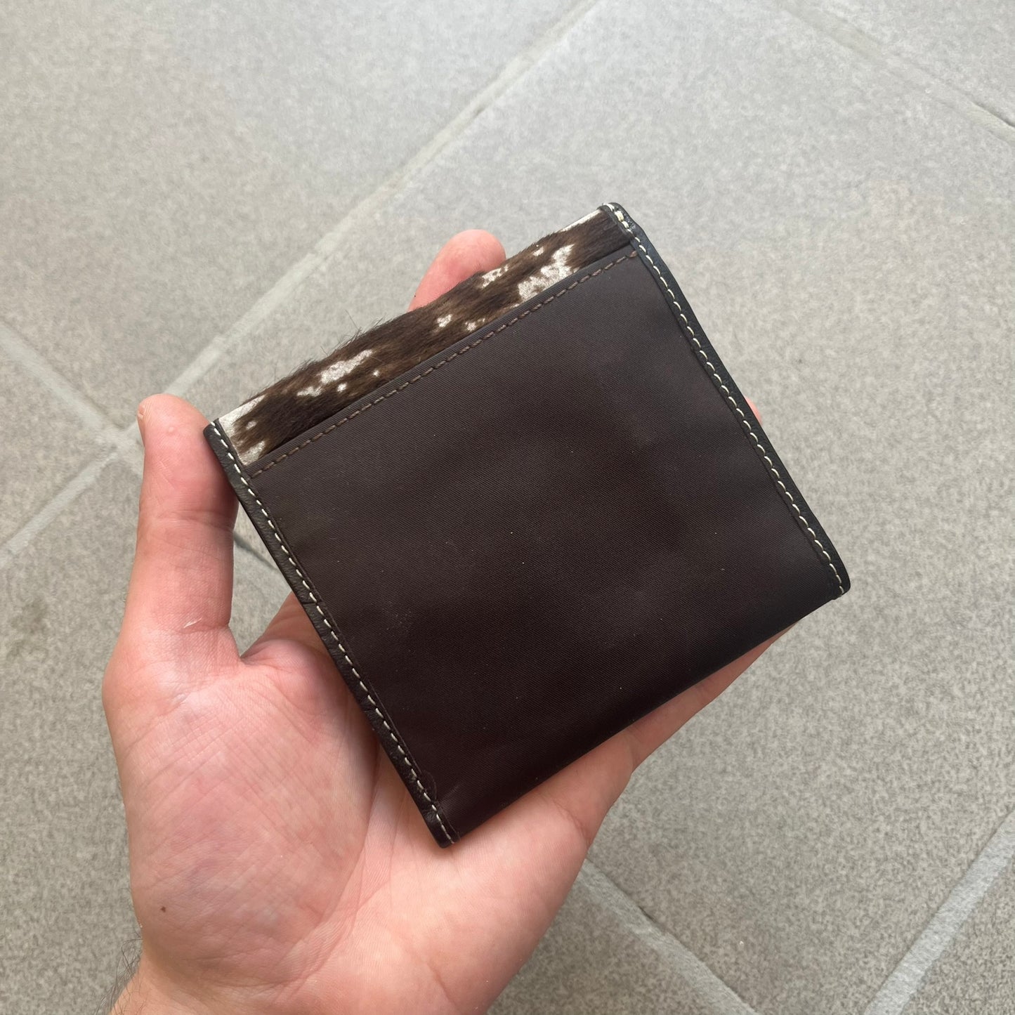 Dolce & Gabbana 2000s Cow-Print Wallet features a brown leather construction with a cow-print pony hair-style panel on the front exterior. A leather patch with a D&G logo is placed over the patterned section. The interior includes multiple compartments, card slots, a top compartment, and a zipped pocket for storage. Made in Italy.
