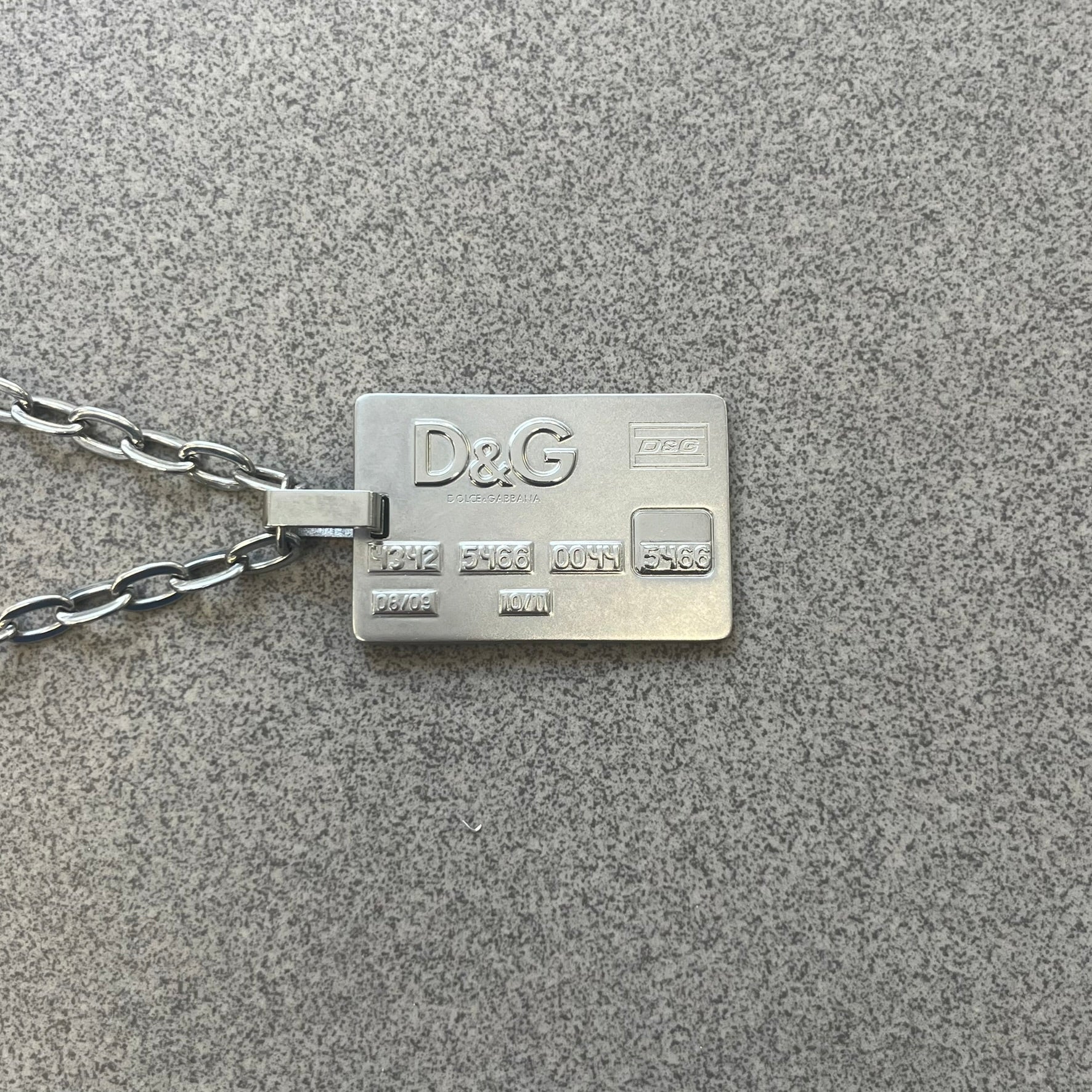 Dolce &amp; Gabbana 2000s Credit Card Necklace (DJ0772) is a rare stainless steel necklace featuring a credit card-shaped pendant. The pendant includes a large raised D&amp;G logo, an engraved D&amp;G logo, and embossed numbers inspired by a real credit card. Finished with a polished metal surface, the necklace comes complete with its original tags and box.