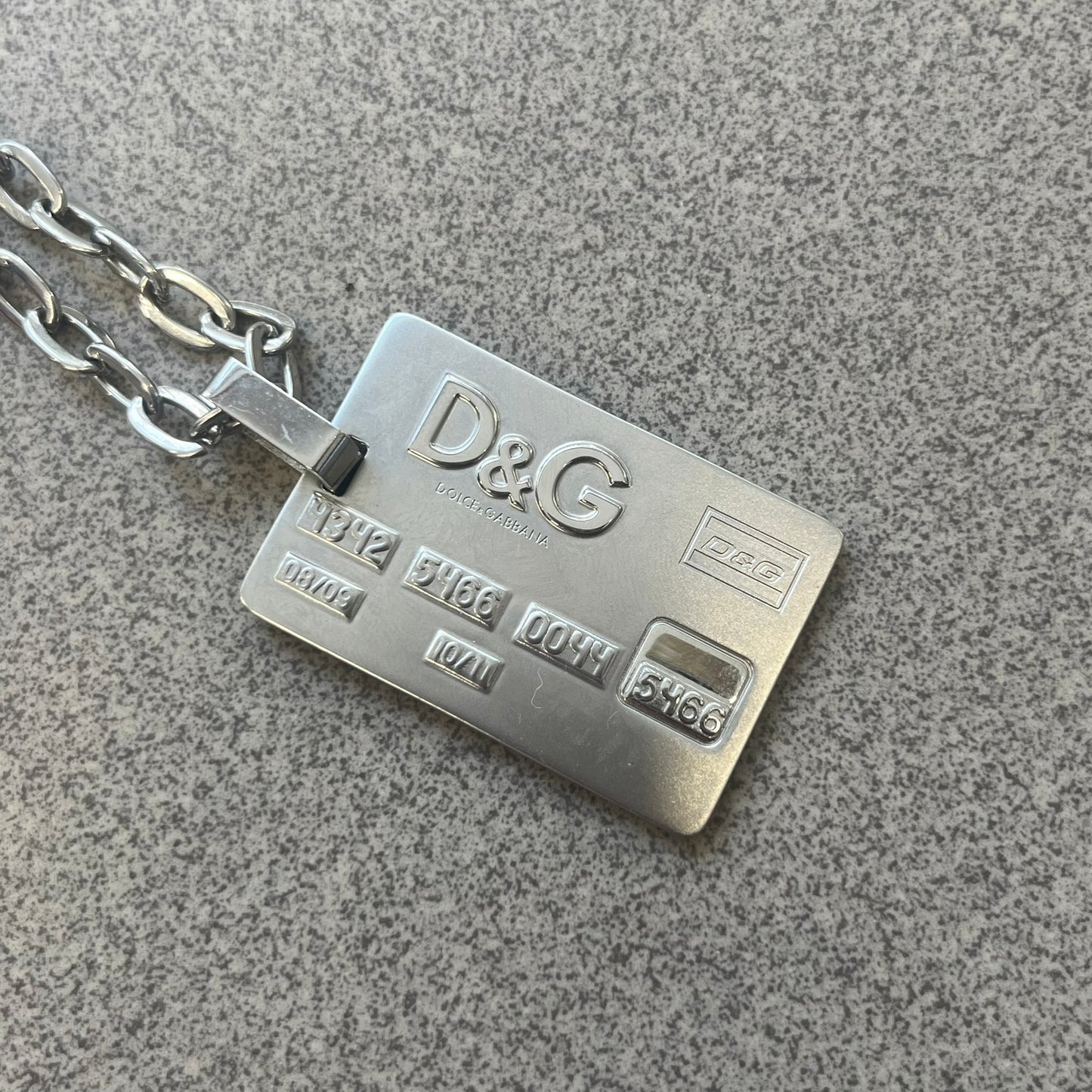 Dolce &amp; Gabbana 2000s Credit Card Necklace (DJ0772) is a rare stainless steel necklace featuring a credit card-shaped pendant. The pendant includes a large raised D&amp;G logo, an engraved D&amp;G logo, and embossed numbers inspired by a real credit card. Finished with a polished metal surface, the necklace comes complete with its original tags and box.