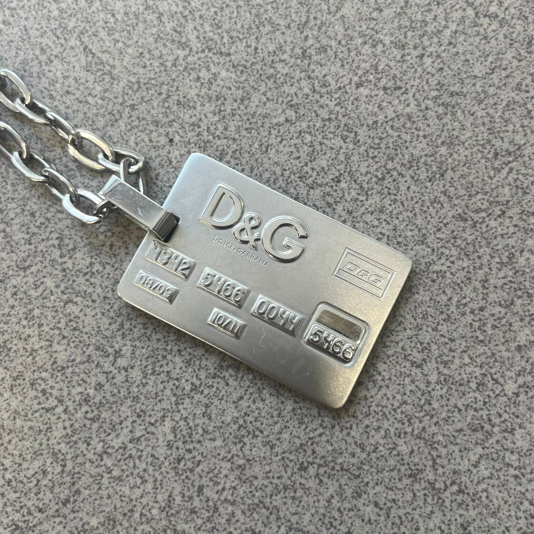 Dolce &amp; Gabbana 2000s Credit Card Necklace (DJ0772) is a rare stainless steel necklace featuring a credit card-shaped pendant. The pendant includes a large raised D&amp;G logo, an engraved D&amp;G logo, and embossed numbers inspired by a real credit card. Finished with a polished metal surface, the necklace comes complete with its original tags and box.