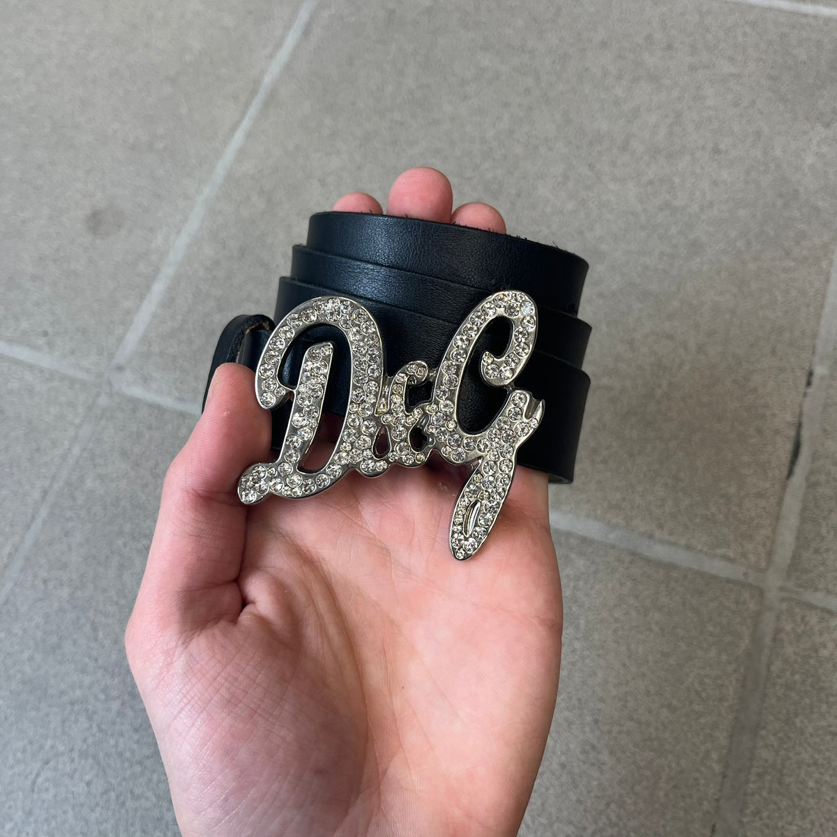 Dolce &amp; Gabbana 2000s Black Crystal Buckle Belt features a black leather strap with a stainless steel D&amp;G logo buckle. The buckle has a decorative italic-style font and is fully covered with crystal/rhinestone detailing on the front. Made in Italy.