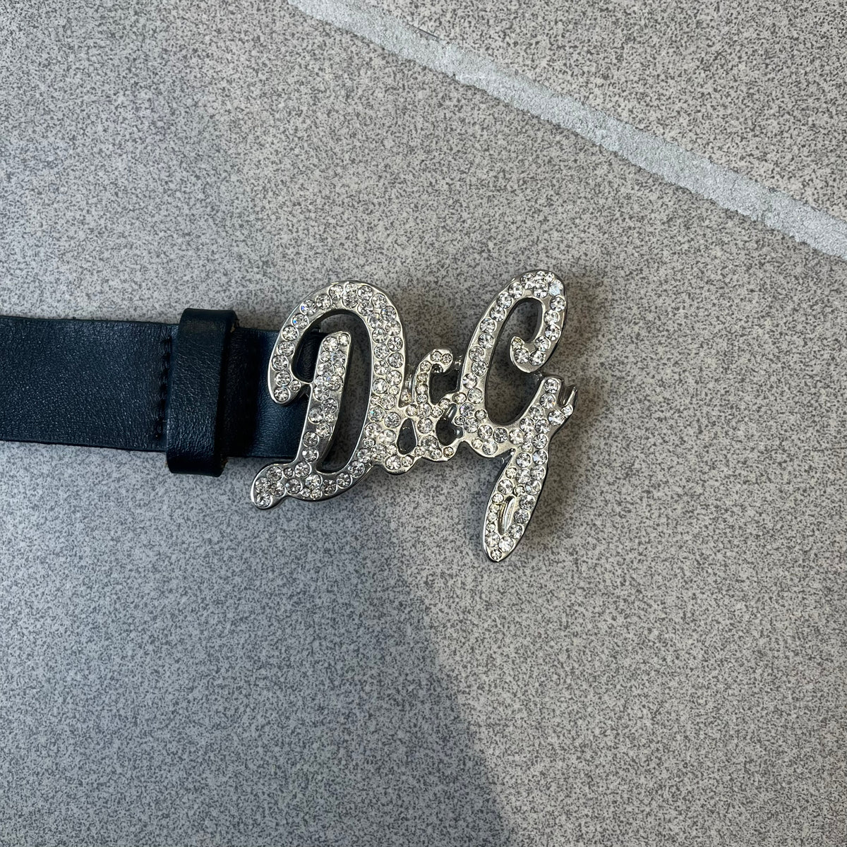 Dolce &amp; Gabbana 2000s Black Crystal Buckle Belt features a black leather strap with a stainless steel D&amp;G logo buckle. The buckle has a decorative italic-style font and is fully covered with crystal/rhinestone detailing on the front. Made in Italy.