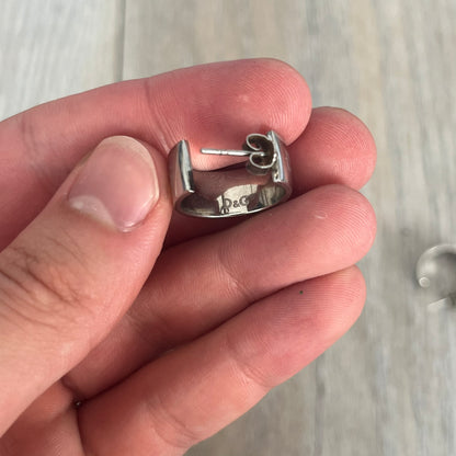 Dolce &amp; Gabbana DJ0404 D&amp;G Logo Earrings are a pair of 2000s silver-tone hoop earrings with a wide profile. The earrings feature an engraved repeating D&amp;G logo around the outer surface. They have a hinged hoop design with a straight post closure and a polished metal finish.