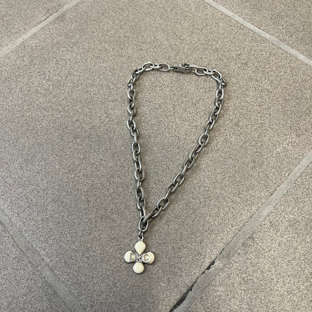 Dolce &amp; Gabbana DJ0879 Four-Leaf Clover Necklace is a stainless steel necklace with a clover pendant that has a stainless steel back and a 3D light beige/cream enamel front. A raised steel D&amp;G logo sits on the front. The oval-link chain alternates between smooth and textured links
