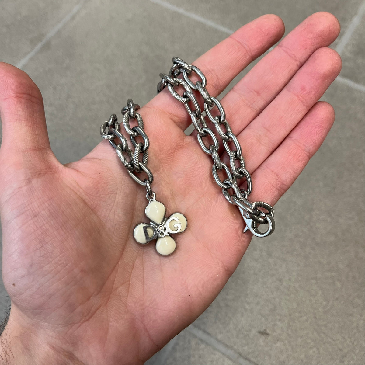 Dolce &amp; Gabbana DJ0879 Four-Leaf Clover Necklace is a stainless steel necklace with a clover pendant that has a stainless steel back and a 3D light beige/cream enamel front. A raised steel D&amp;G logo sits on the front. The oval-link chain alternates between smooth and textured links