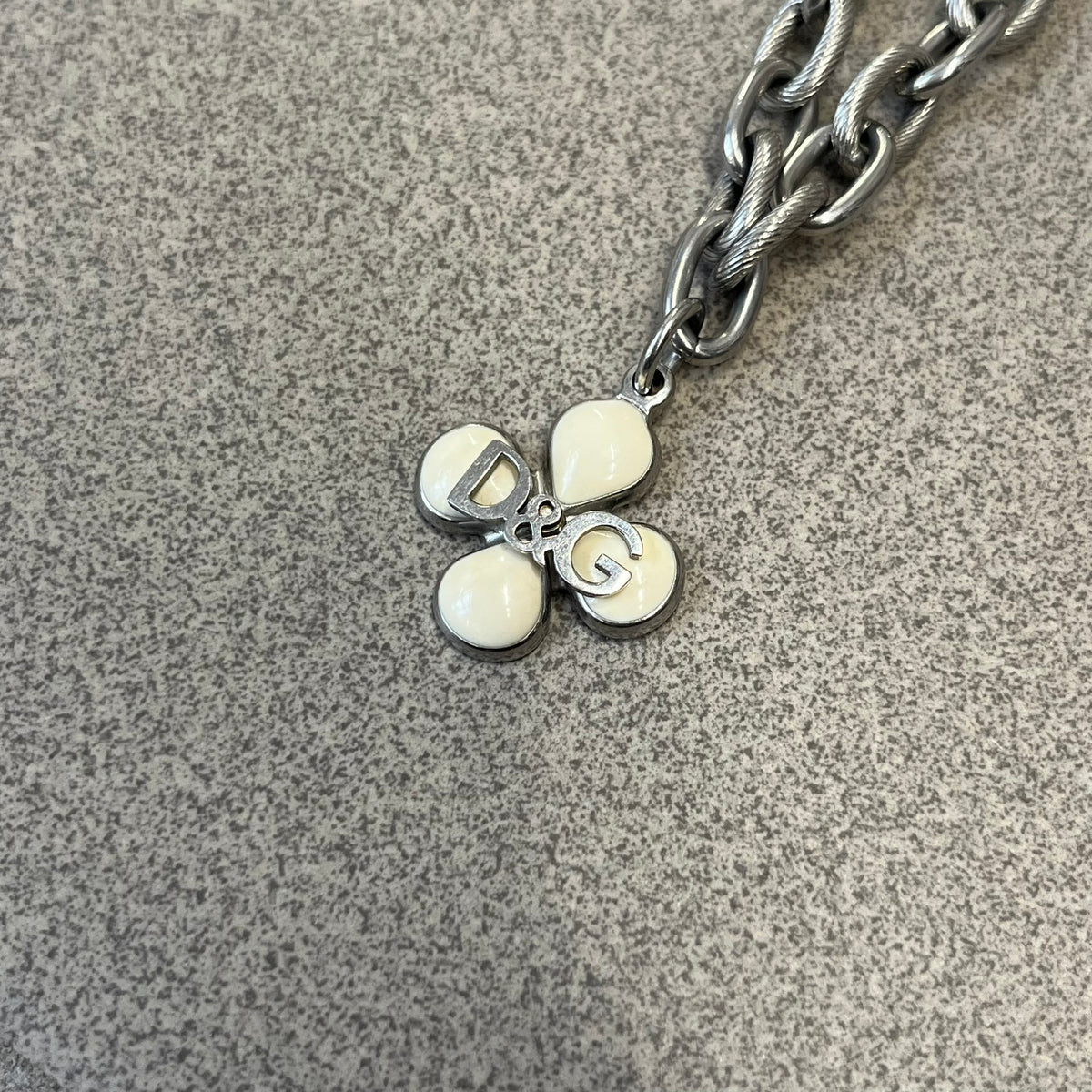 Dolce &amp; Gabbana DJ0879 Four-Leaf Clover Necklace is a stainless steel necklace with a clover pendant that has a stainless steel back and a 3D light beige/cream enamel front. A raised steel D&amp;G logo sits on the front. The oval-link chain alternates between smooth and textured links