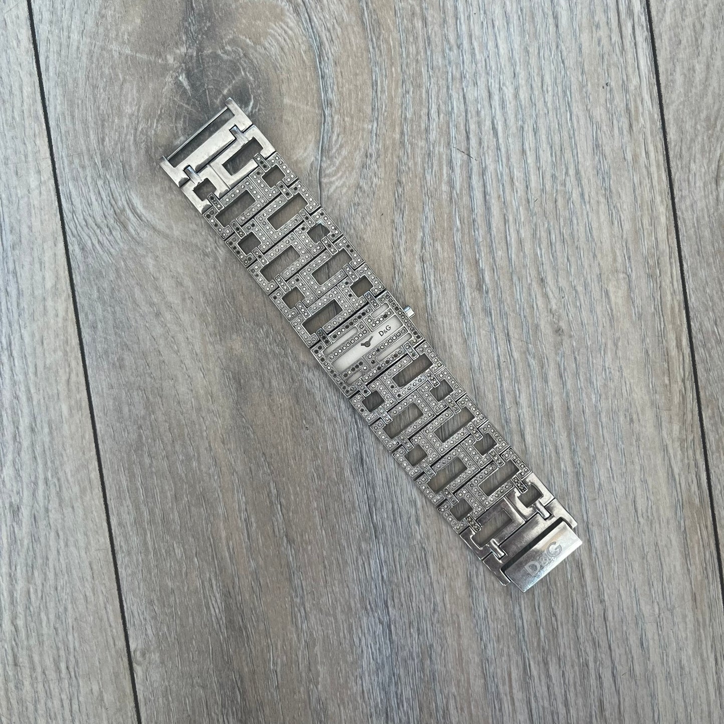 Dolce & Gabbana 2000s Geometric Analog Watch is a silver-tone watch with a matching bracelet and case that maintain the same width throughout. The slim rectangular white dial features small hands and a subtle D&G logo. The case and bracelet are set with rhinestones, while the bracelet has an irregular pattern of cut-out rectangular and square links. The watch comes with its original box, papers, and tag.
