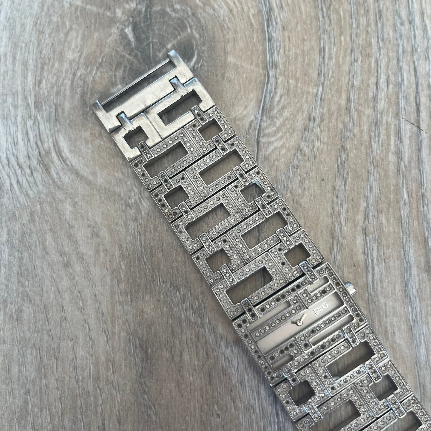 Dolce & Gabbana 2000s Geometric Analog Watch is a silver-tone watch with a matching bracelet and case that maintain the same width throughout. The slim rectangular white dial features small hands and a subtle D&G logo. The case and bracelet are set with rhinestones, while the bracelet has an irregular pattern of cut-out rectangular and square links. The watch comes with its original box, papers, and tag.
