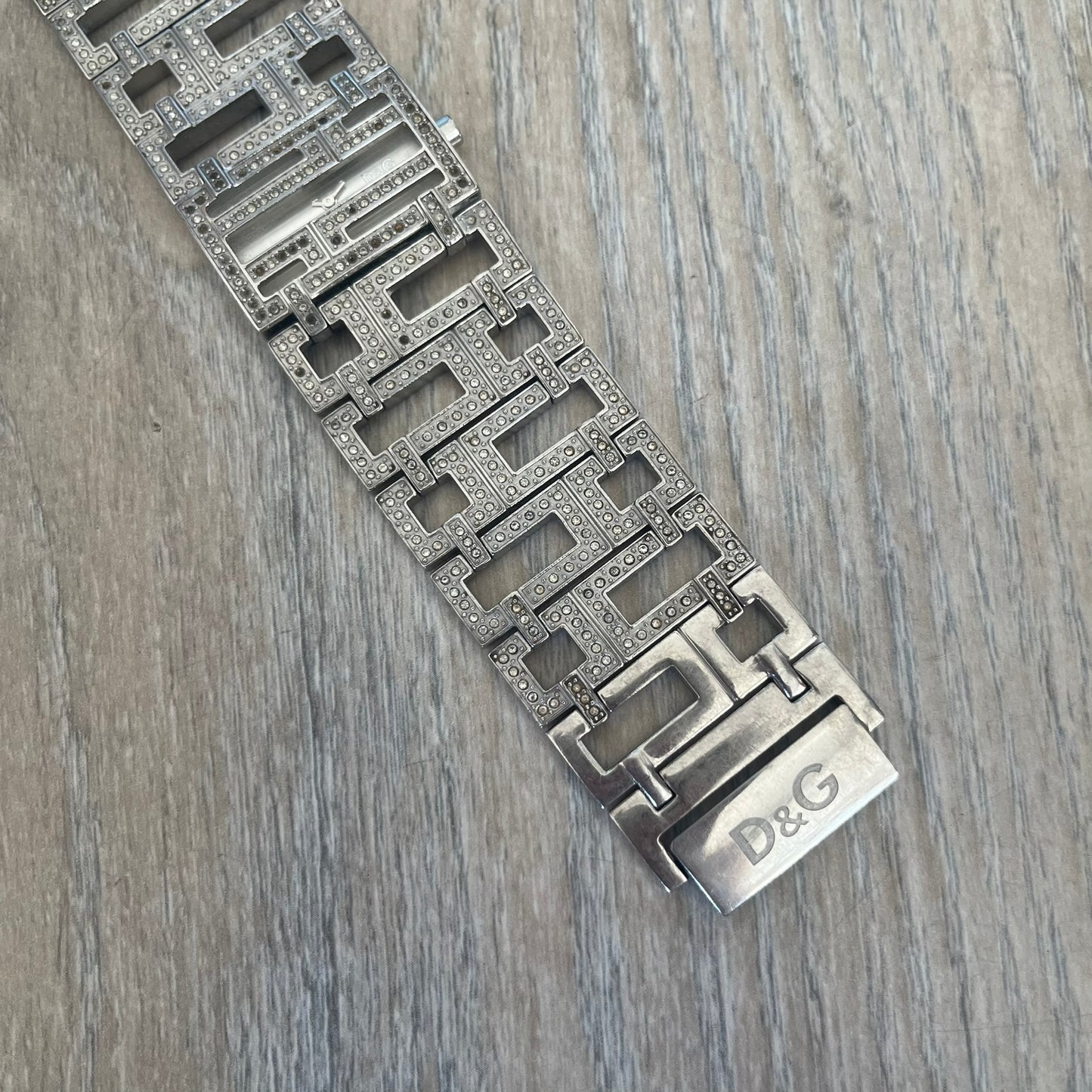 Dolce & Gabbana 2000s Geometric Analog Watch is a silver-tone watch with a matching bracelet and case that maintain the same width throughout. The slim rectangular white dial features small hands and a subtle D&G logo. The case and bracelet are set with rhinestones, while the bracelet has an irregular pattern of cut-out rectangular and square links. The watch comes with its original box, papers, and tag.
