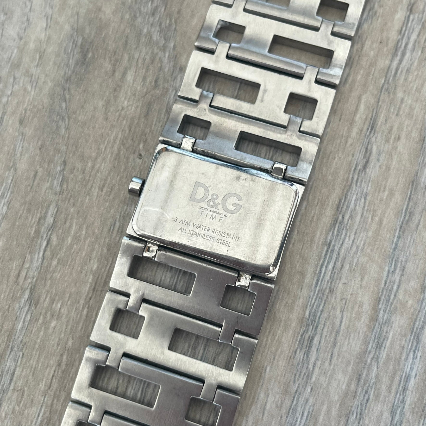 Dolce & Gabbana 2000s Geometric Analog Watch is a silver-tone watch with a matching bracelet and case that maintain the same width throughout. The slim rectangular white dial features small hands and a subtle D&G logo. The case and bracelet are set with rhinestones, while the bracelet has an irregular pattern of cut-out rectangular and square links. The watch comes with its original box, papers, and tag.

