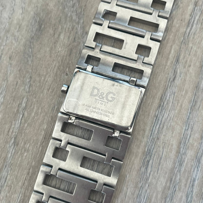 Dolce & Gabbana 2000s Geometric Analog Watch is a silver-tone watch with a matching bracelet and case that maintain the same width throughout. The slim rectangular white dial features small hands and a subtle D&G logo. The case and bracelet are set with rhinestones, while the bracelet has an irregular pattern of cut-out rectangular and square links. The watch comes with its original box, papers, and tag.
