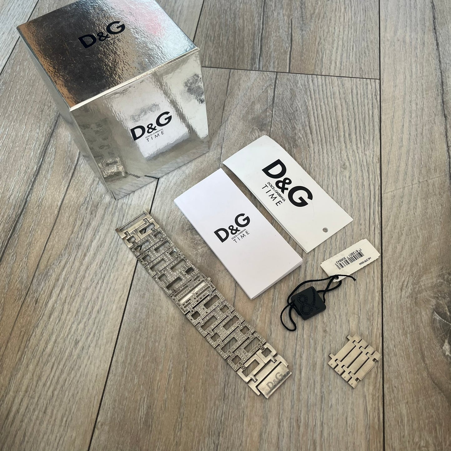Dolce & Gabbana 2000s Geometric Analog Watch is a silver-tone watch with a matching bracelet and case that maintain the same width throughout. The slim rectangular white dial features small hands and a subtle D&G logo. The case and bracelet are set with rhinestones, while the bracelet has an irregular pattern of cut-out rectangular and square links. The watch comes with its original box, papers, and tag.
