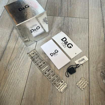 Dolce & Gabbana 2000s Geometric Analog Watch is a silver-tone watch with a matching bracelet and case that maintain the same width throughout. The slim rectangular white dial features small hands and a subtle D&G logo. The case and bracelet are set with rhinestones, while the bracelet has an irregular pattern of cut-out rectangular and square links. The watch comes with its original box, papers, and tag.

