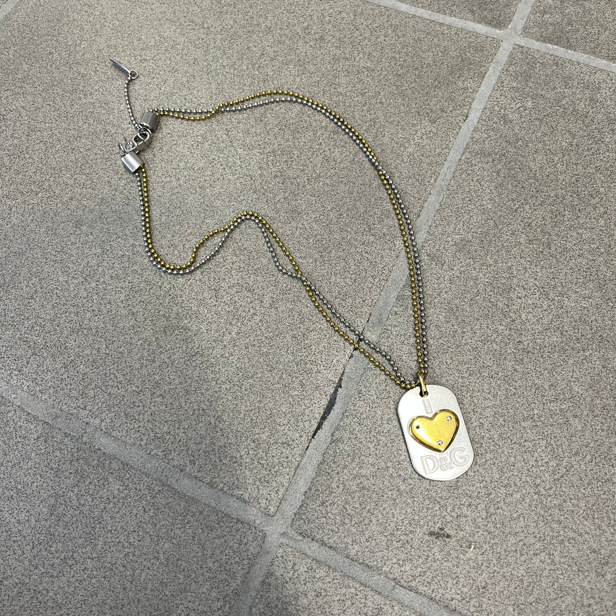 Dolce &amp; Gabbana 2000's Gold Heart DJ0637 Necklace features a stainless steel ball &nbsp;chain, one in silver-tone, the second in gold-tone, with a dog tag-style pendant. The pendant is engraved with a subtle "I" above a gold-tone heart secured by three visible screws, with "D&amp;G" engraved below to form the "I ❤️ D&amp;G" design.&nbsp;