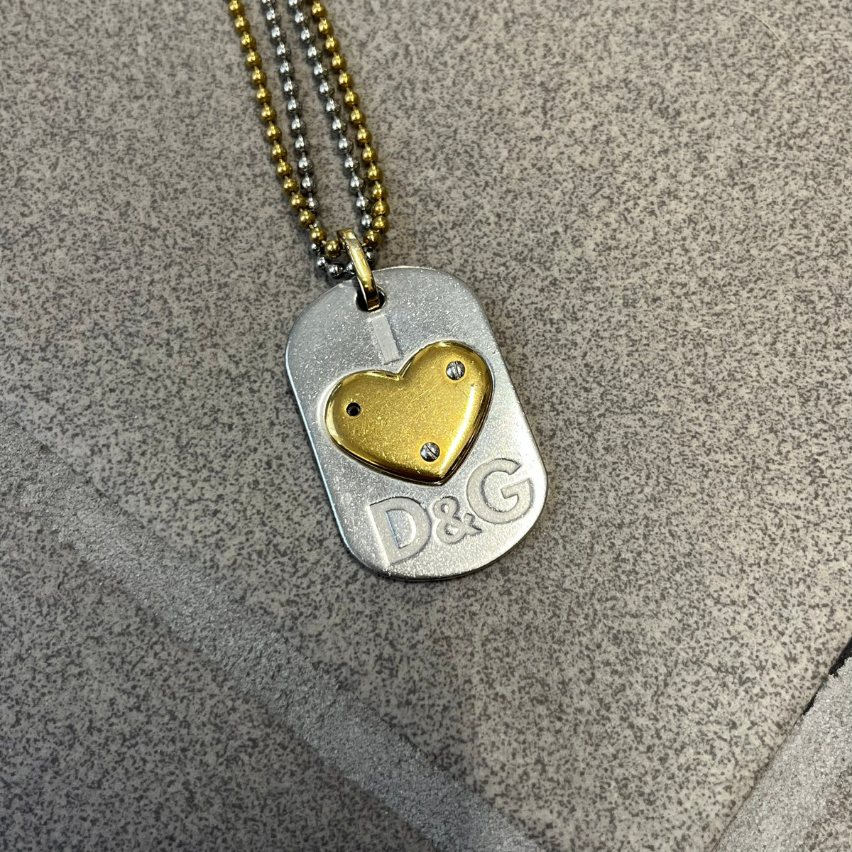 Dolce &amp; Gabbana 2000's Gold Heart DJ0637 Necklace features a stainless steel ball &nbsp;chain, one in silver-tone, the second in gold-tone, with a dog tag-style pendant. The pendant is engraved with a subtle "I" above a gold-tone heart secured by three visible screws, with "D&amp;G" engraved below to form the "I ❤️ D&amp;G" design.&nbsp;