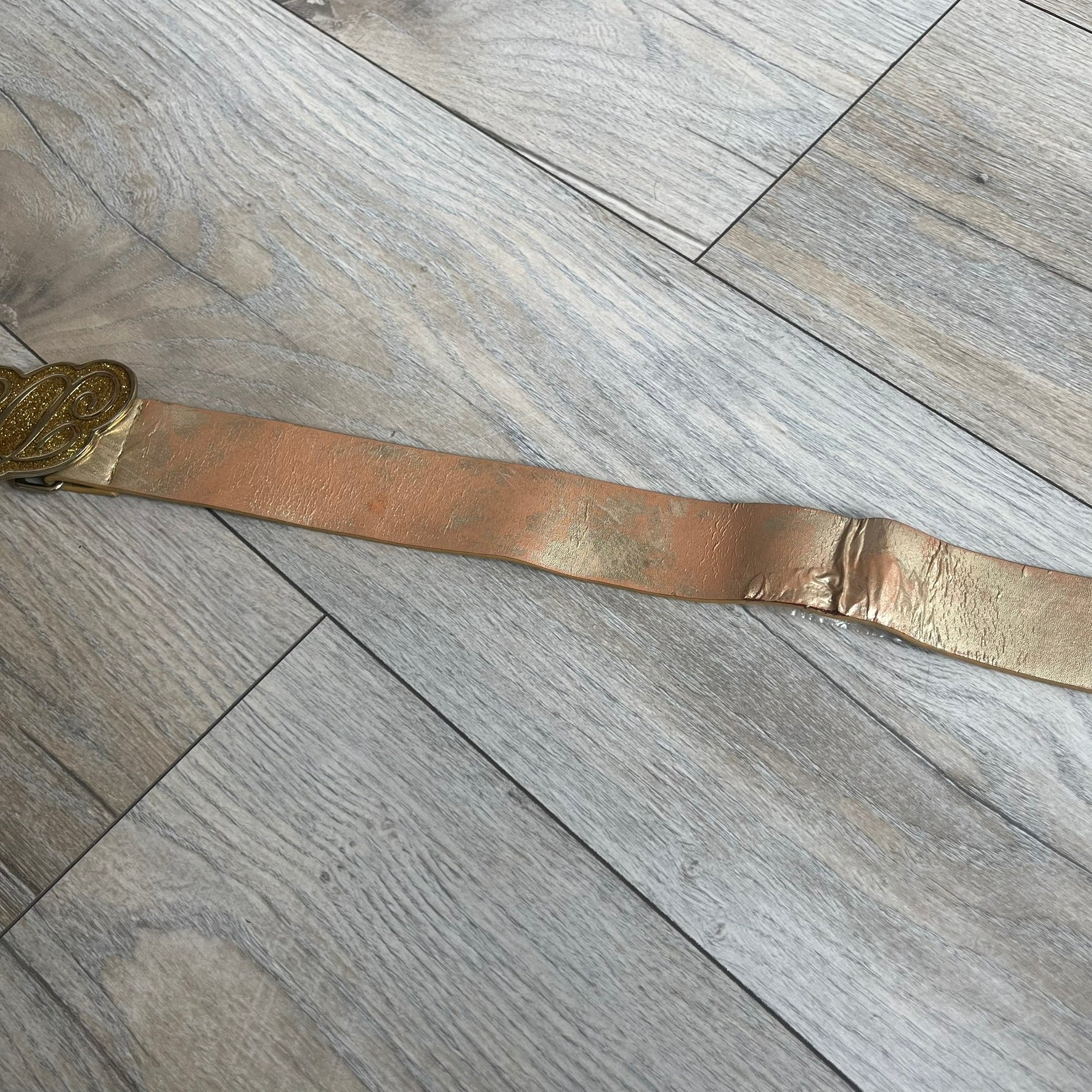The Dolce &amp; Gabbana 2000’s Gold Tone D&amp;G Logo Belt is made from metallic gold leather and features a large D&amp;G logo buckle shaped directly as the brand’s initials. The buckle uses a cursive D&amp;G logo set over a glittery gold background, framed by a solid gold-tone metal outline. The entire buckle and belt maintain a consistent gold color scheme typical of Dolce &amp; Gabbana accessories from the 2000s.