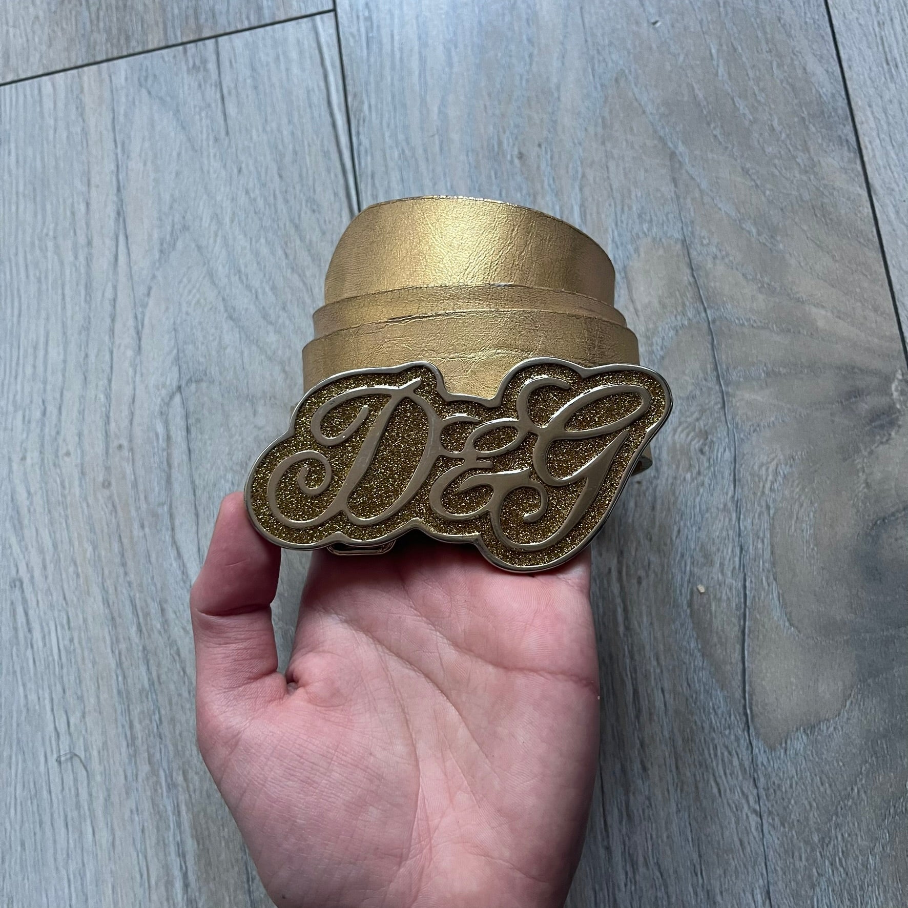 The Dolce & Gabbana 2000’s Gold Tone D&G Logo Belt is made from metallic gold leather and features a large D&G logo buckle shaped directly as the brand’s initials. The buckle uses a cursive D&G logo set over a glittery gold background, framed by a solid gold-tone metal outline. The entire buckle and belt maintain a consistent gold color scheme typical of Dolce & Gabbana accessories from the 2000s.