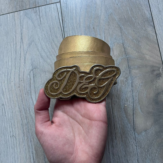 The Dolce & Gabbana 2000’s Gold Tone D&G Logo Belt is made from metallic gold leather and features a large D&G logo buckle shaped directly as the brand’s initials. The buckle uses a cursive D&G logo set over a glittery gold background, framed by a solid gold-tone metal outline. The entire buckle and belt maintain a consistent gold color scheme typical of Dolce & Gabbana accessories from the 2000s.