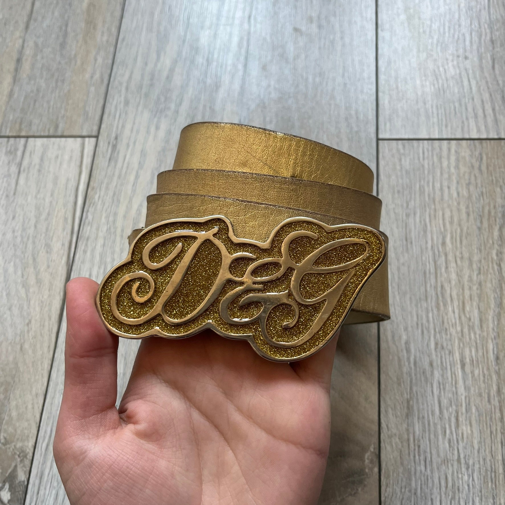 The Dolce &amp; Gabbana 2000’s Gold Tone D&amp;G Logo Belt is made from metallic gold leather and features a large D&amp;G logo buckle shaped directly as the brand’s initials. The buckle uses a cursive D&amp;G logo set over a glittery gold background, framed by a solid gold-tone metal outline. The entire buckle and belt maintain a consistent gold color scheme typical of Dolce &amp; Gabbana accessories from the 2000s.
