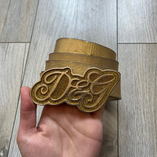 The Dolce &amp; Gabbana 2000’s Gold Tone D&amp;G Logo Belt is made from metallic gold leather and features a large D&amp;G logo buckle shaped directly as the brand’s initials. The buckle uses a cursive D&amp;G logo set over a glittery gold background, framed by a solid gold-tone metal outline. The entire buckle and belt maintain a consistent gold color scheme typical of Dolce &amp; Gabbana accessories from the 2000s.