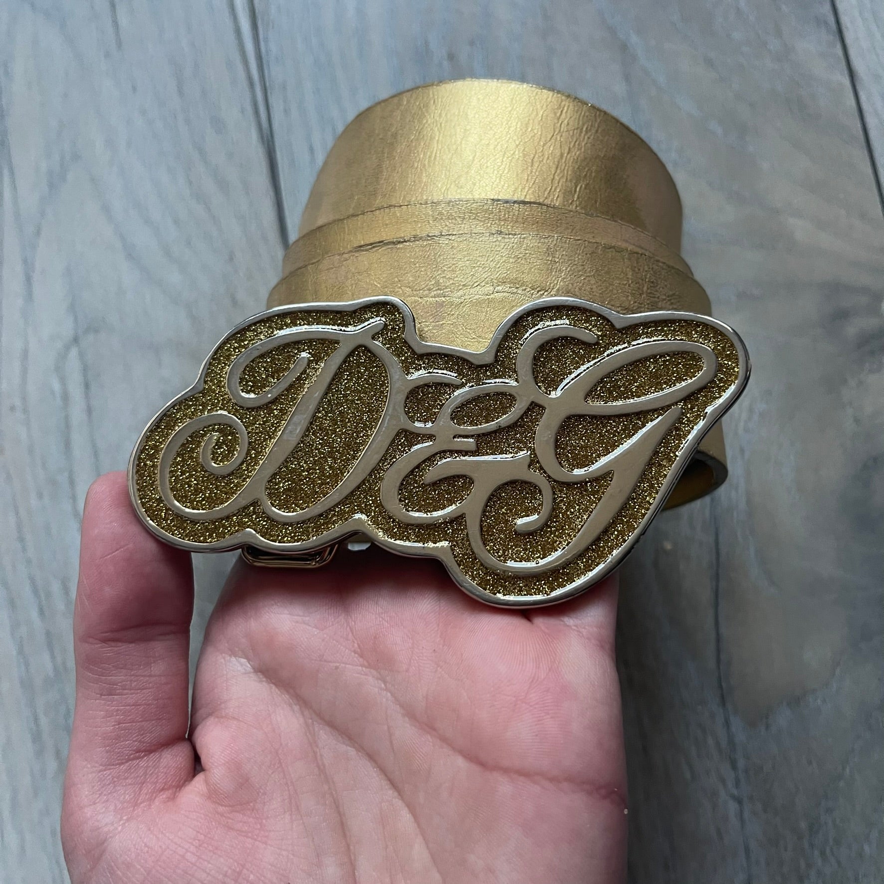 The Dolce & Gabbana 2000’s Gold Tone D&G Logo Belt is made from metallic gold leather and features a large D&G logo buckle shaped directly as the brand’s initials. The buckle uses a cursive D&G logo set over a glittery gold background, framed by a solid gold-tone metal outline. The entire buckle and belt maintain a consistent gold color scheme typical of Dolce & Gabbana accessories from the 2000s.