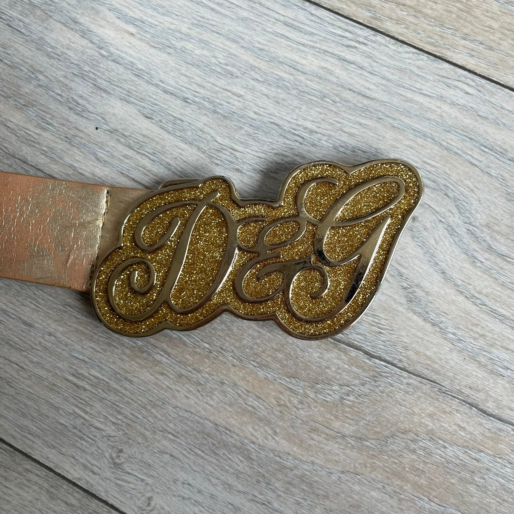 The Dolce &amp; Gabbana 2000’s Gold Tone D&amp;G Logo Belt is made from metallic gold leather and features a large D&amp;G logo buckle shaped directly as the brand’s initials. The buckle uses a cursive D&amp;G logo set over a glittery gold background, framed by a solid gold-tone metal outline. The entire buckle and belt maintain a consistent gold color scheme typical of Dolce &amp; Gabbana accessories from the 2000s.