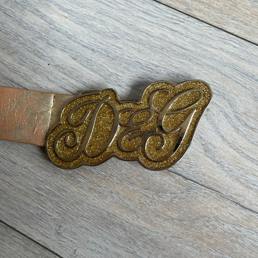 The Dolce &amp; Gabbana 2000’s Gold Tone D&amp;G Logo Belt is made from metallic gold leather and features a large D&amp;G logo buckle shaped directly as the brand’s initials. The buckle uses a cursive D&amp;G logo set over a glittery gold background, framed by a solid gold-tone metal outline. The entire buckle and belt maintain a consistent gold color scheme typical of Dolce &amp; Gabbana accessories from the 2000s.
